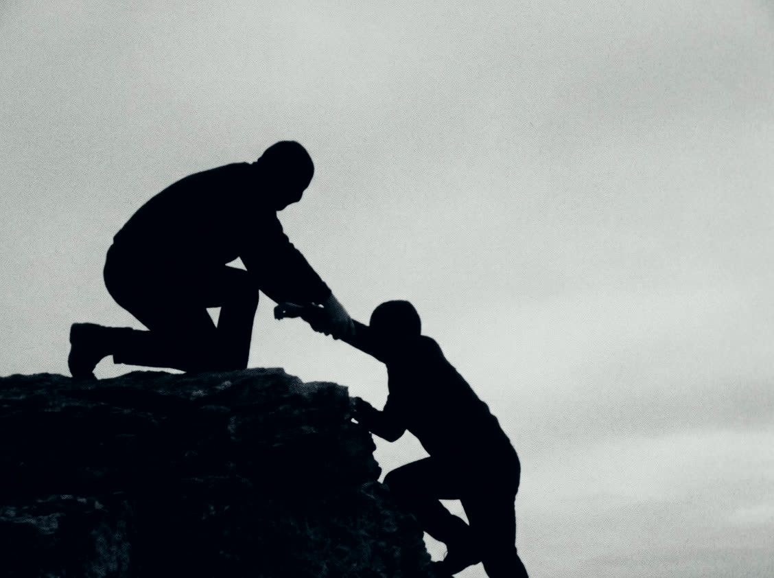 Two silhouettes on a rock shelf. One pulls the other up.