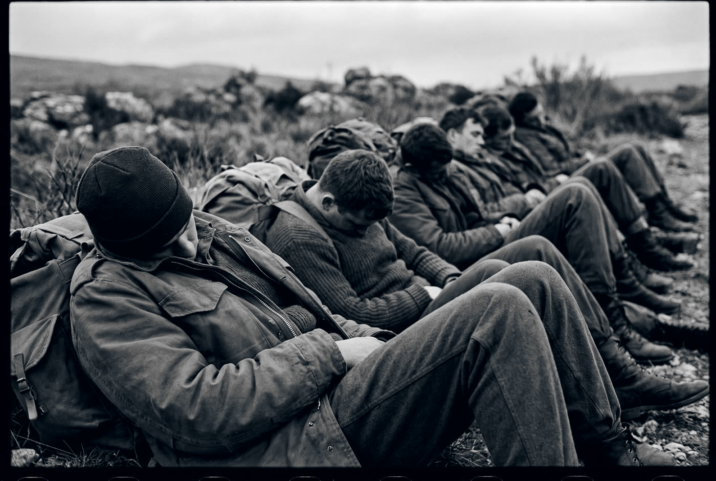 Spent men asleep sitting upright against their rucks in the dawn mist.