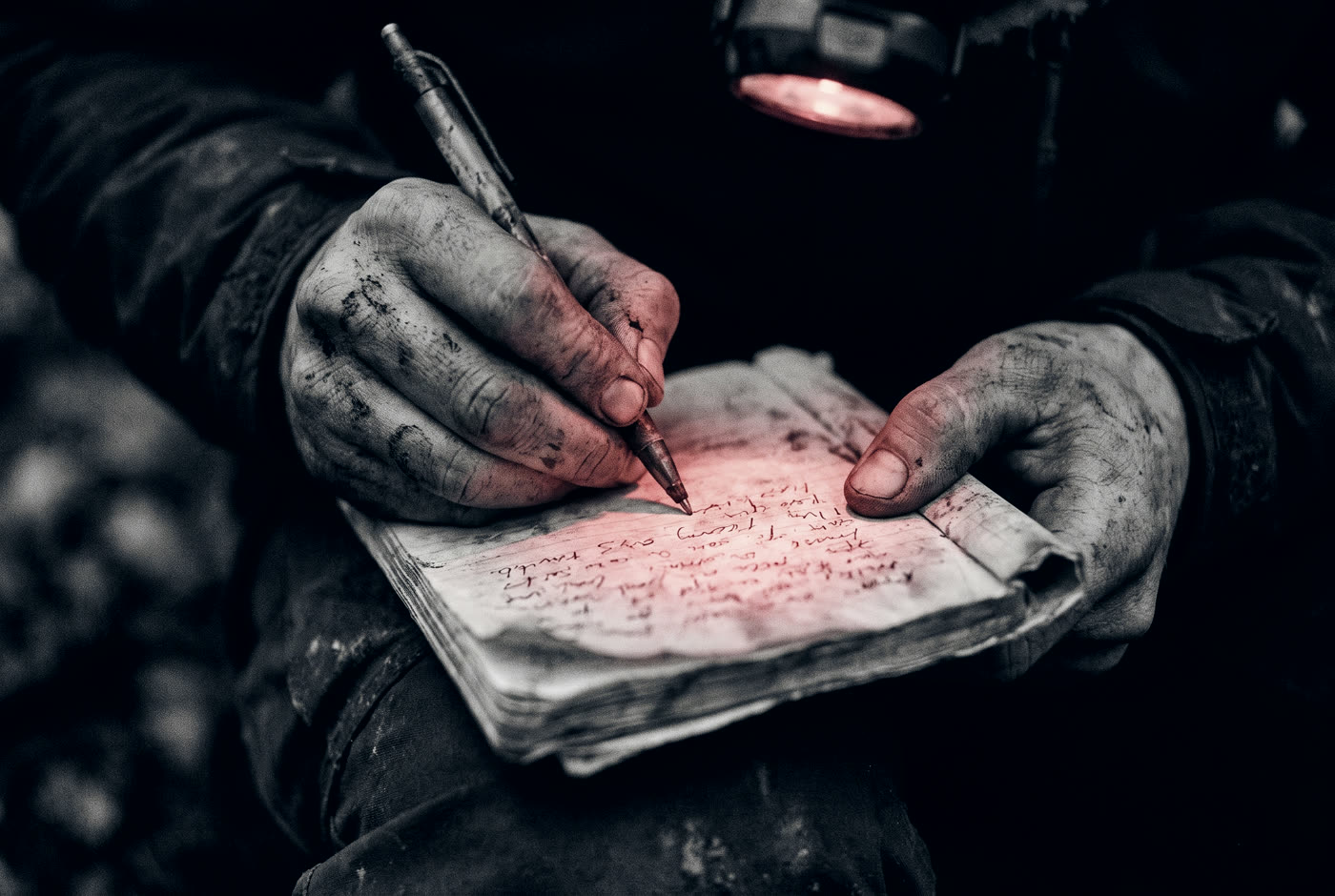 Mud-caked hands writing in a journal by the light of a red headlamp.