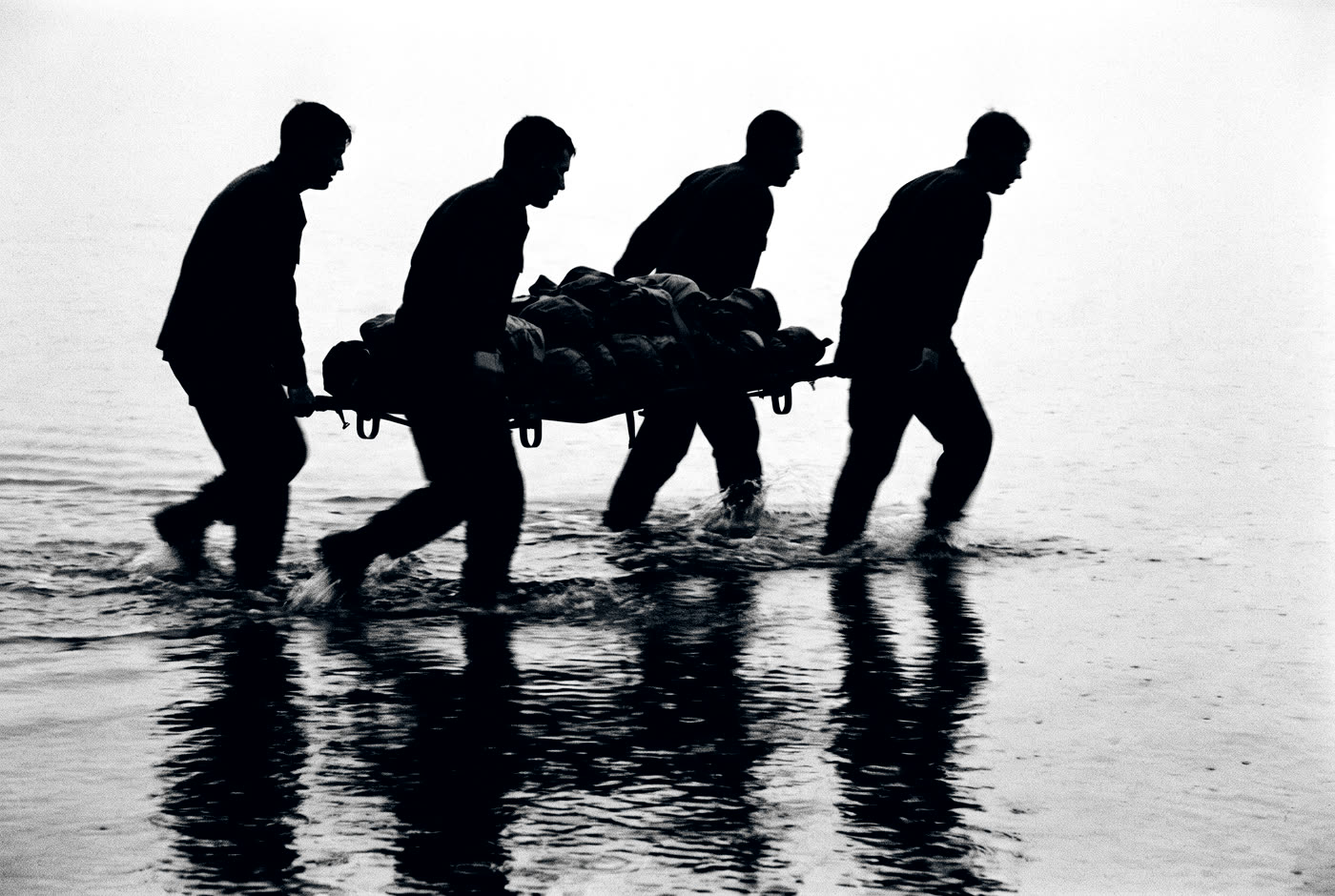 Four men carrying a loaded litter through shallow water at first light, in silhouette.