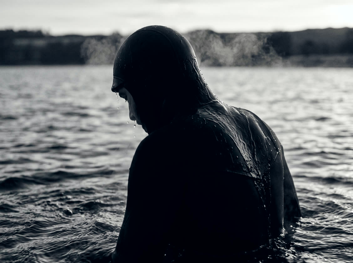 A figure surfacing from black water, head bowed, breath steaming in cold air.